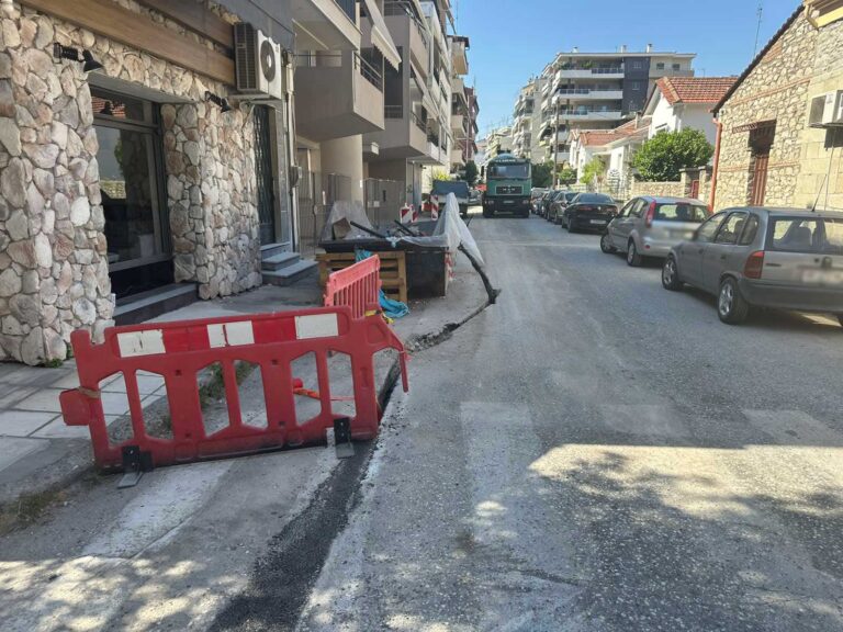 Κατασκευαστικές εργασίες σε δρόμο των Τρικάλων με κόκκινα προστατευτικά και κάδο μπαζών να εμποδίζει την ευθεία χάραξη της τομής.