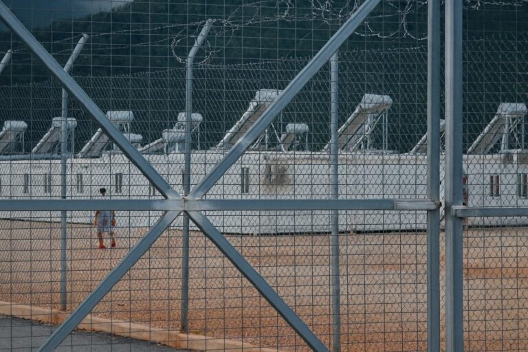 Εγκαταστάσεις δομής φιλοξενίας πίσω από ψηλό φράχτη με συρματόπλεγμα, με εμφανή τα κοντέινερ και έναν άνθρωπο να περπατά στον περίβολο — υπονοείται περιορισμένη πρόσβαση και κλειστός χαρακτήρας της δομής.