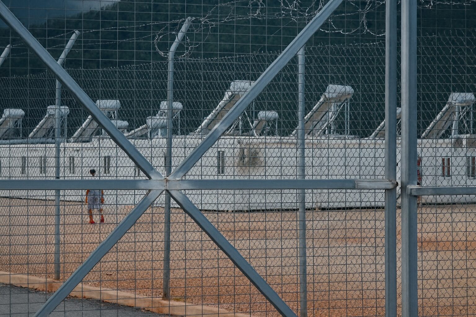 Εγκαταστάσεις δομής φιλοξενίας πίσω από ψηλό φράχτη με συρματόπλεγμα, με εμφανή τα κοντέινερ και έναν άνθρωπο να περπατά στον περίβολο — υπονοείται περιορισμένη πρόσβαση και κλειστός χαρακτήρας της δομής.