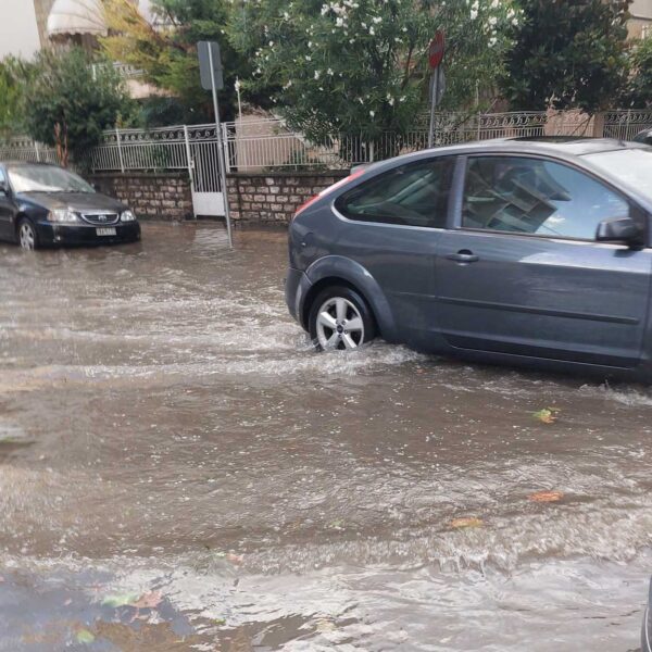 Αυτοκίνητα περνούν μέσα από πλημμυρισμένο δρόμο στη διασταύρωση Απόλλωνος και Πολυκάρπου στα Τρίκαλα, μετά από βροχόπτωση και φραγμένα φρεάτια.