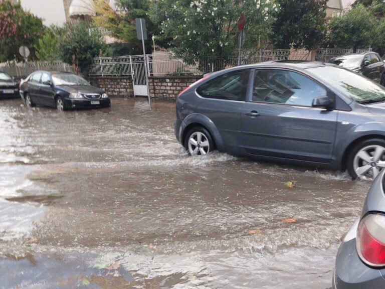 Αυτοκίνητα περνούν μέσα από πλημμυρισμένο δρόμο στη διασταύρωση Απόλλωνος και Πολυκάρπου στα Τρίκαλα, μετά από βροχόπτωση και φραγμένα φρεάτια.