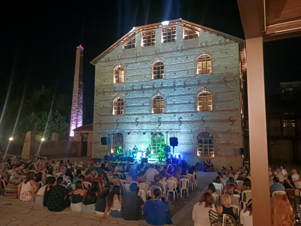 Μουσική βραδιά στον Μύλο Ματσόπουλου με φόντο το φωτισμένο κτίριο και τον κόσμο.
