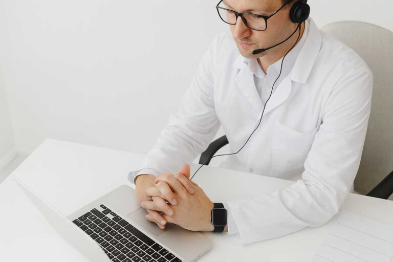 ιατρός με λευκή ιατρική ποδιά και ακουστικό headset εργάζεται σε φορητό υπολογιστή σε σύγχρονο ιατρείο.