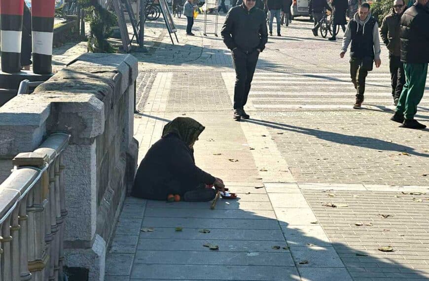 Ηλικιωμένη γυναίκα σε στάση επαιτείας στην κεντρική γέφυρα Τρικάλων, ενώ πολίτες κινούνται στο κέντρο της πόλης τις ημέρες των Χριστουγέννων.