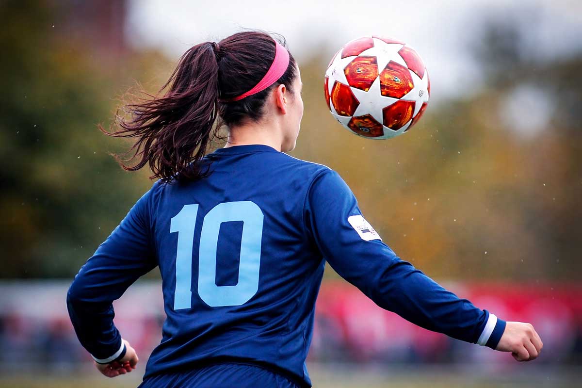 Νεαρή ποδοσφαιρίστρια με μπλε φανέλα και κοτσίδα κάνει κοντρόλ με το κεφάλι σε μπάλα ποδοσφαίρου κατά τη διάρκεια αγώνα.