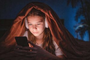 Ανήλικη χρησιμοποιεί κινητό τη νύχτα κάτω από κουβέρτα, φωτισμένη από την οθόνη, συμβολίζοντας τη χρήση social media σε παιδιά