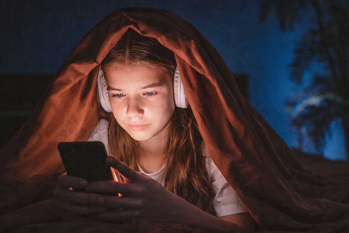 Ανήλικη χρησιμοποιεί κινητό τη νύχτα κάτω από κουβέρτα, φωτισμένη από την οθόνη, συμβολίζοντας τη χρήση social media σε παιδιά